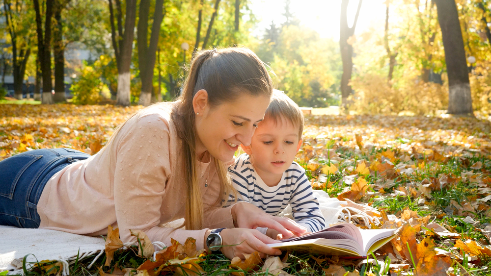 Portrait of young mother telling a story to her little son lying on yellow fallen tree leaves at autumn park.