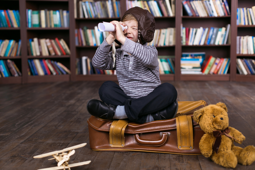 Little boy having fun at room with bookshelf and sitting on suitcase. 