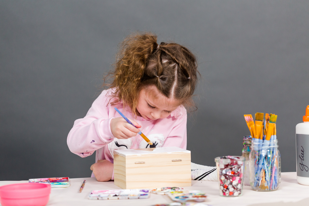 Little girl painting a unicorn on a wood box with acrylic paint.