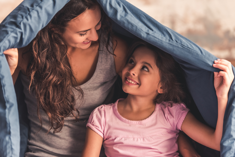 Beautiful mom and daughter are looking at each other and smiling while sitting on bed covered with a duvet