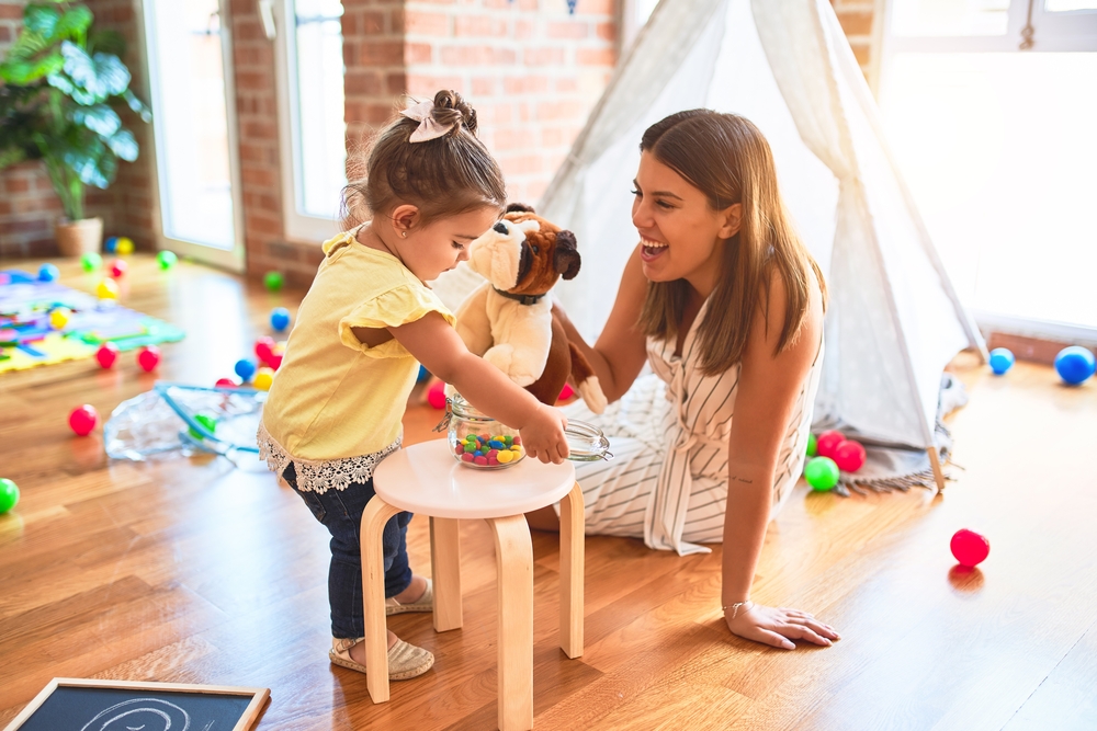 Young beautiful teacher playing with dog doll and toddler holding jar of chocolate balls at kindergarten