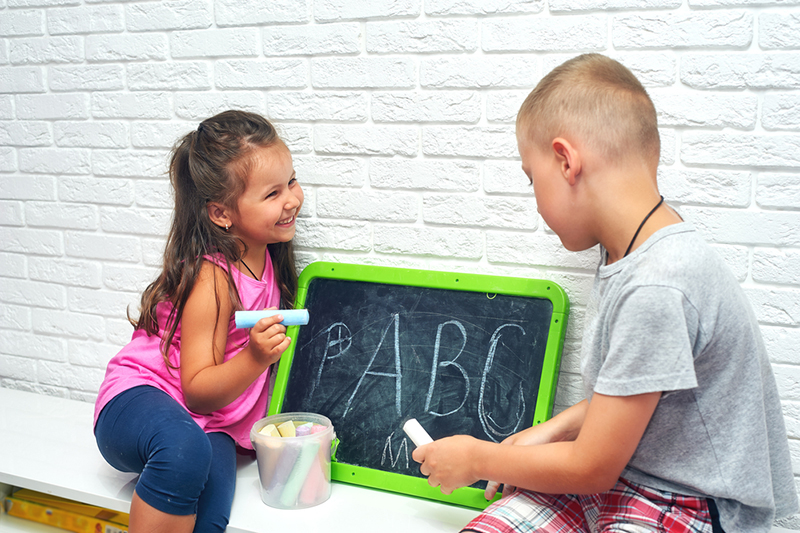 Two kids writing uppercase letters on a chalk board